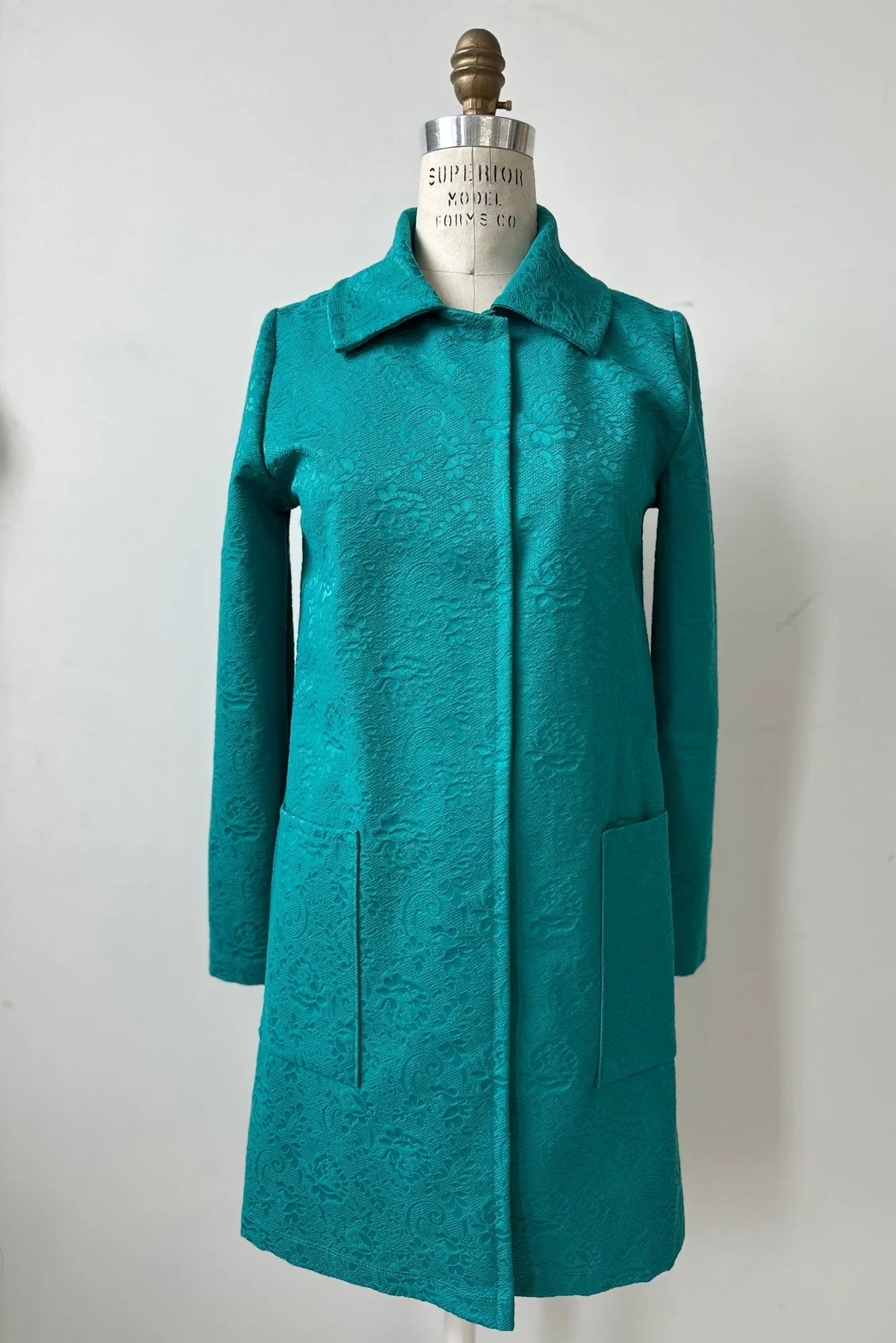The Emmanuelle Coat by Kollontai in Turquoise jacquard fabric is shown on a mannequin in front of a white background. It has a straight cut, classic collar, large patch pockets, and falls just above the knee.