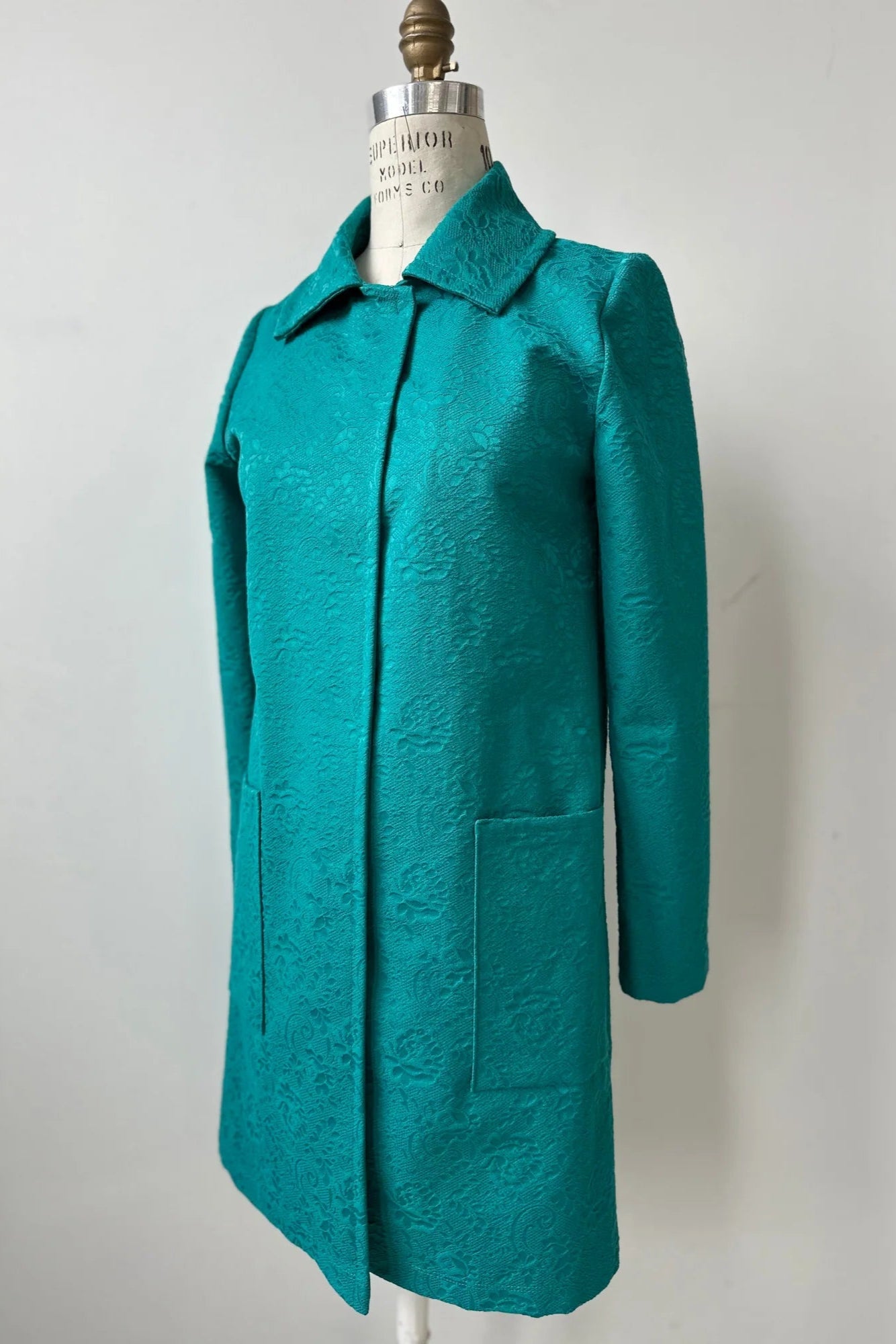 The Emmanuelle Coat by Kollontai in Turquoise jacquard fabric is shown on a mannequin in front of a white background. It has a straight cut, classic collar, large patch pockets, and falls just above the knee.