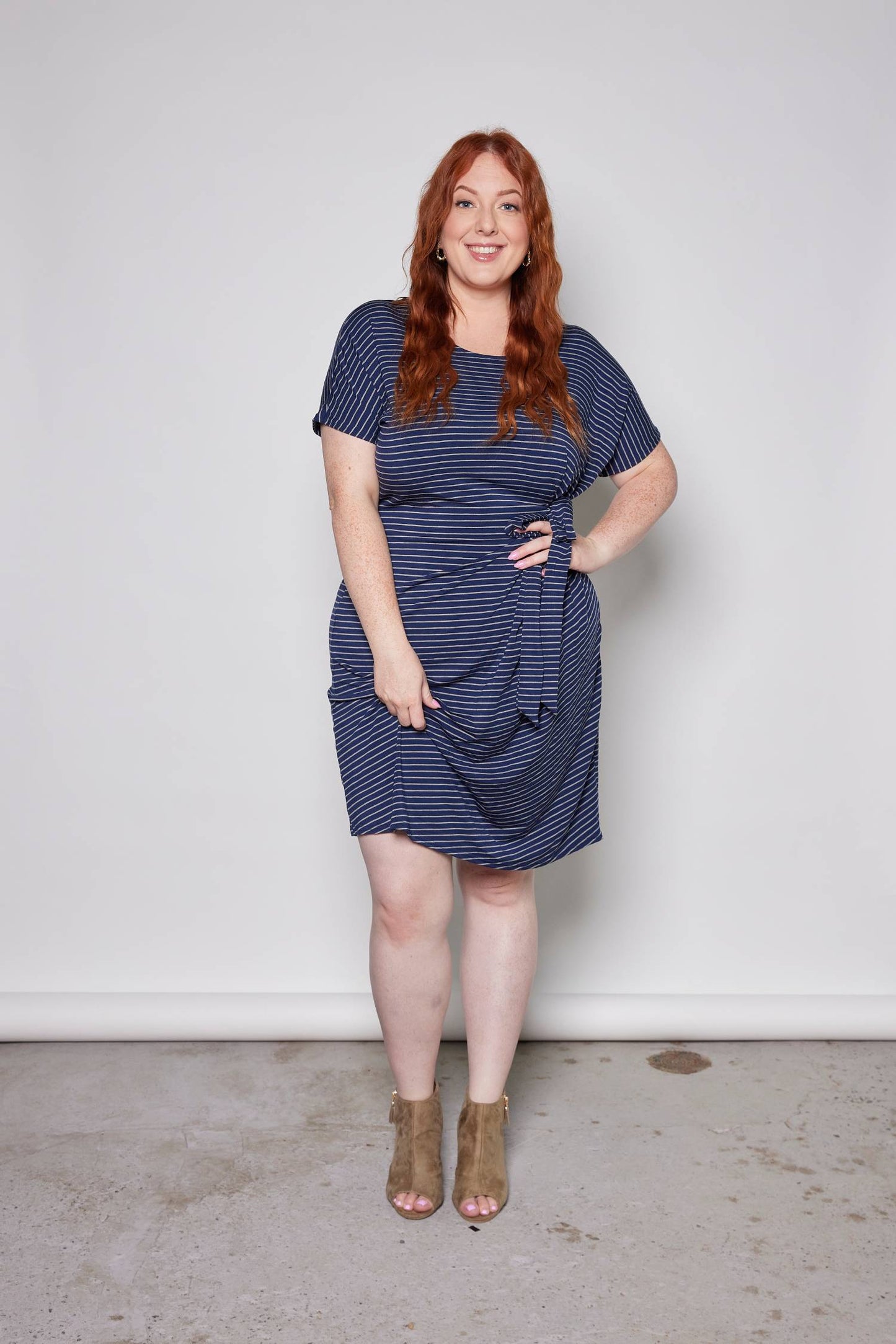 A woman wearing the Joell Dress by Tangente in Blue/Grey, a cap-sleeved dress in striped bamboo with a tie at the waist and an above-the-knee hem. She is standing in front of a white wall.
