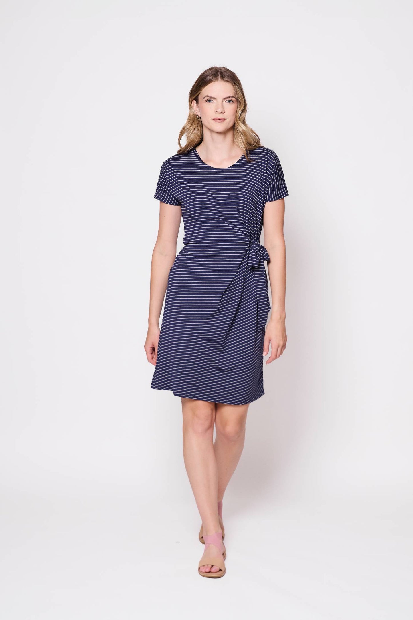 A woman wearing the Joelle Dress by Tangente in Blue/Grey, a cap-sleeved dress in striped bamboo with a tie at the waist and an above-the-knee hem. She is standing in front of a white wall.