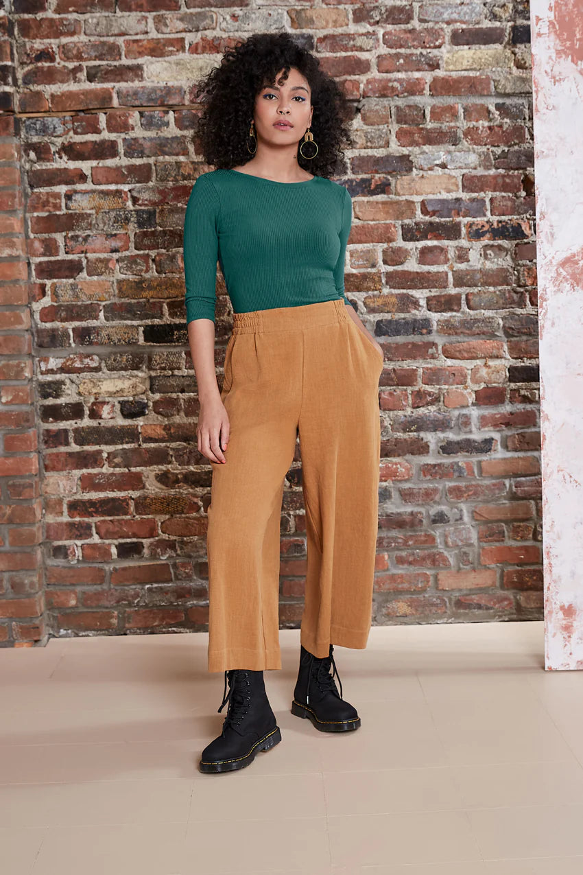 A woman wearing the Kurt Pants by Melow in Honey, featuring wide cropped legs, a flat-front elastic waist band, and slant pockets. She is wearing them with a green top and standing in front of a brick wall.