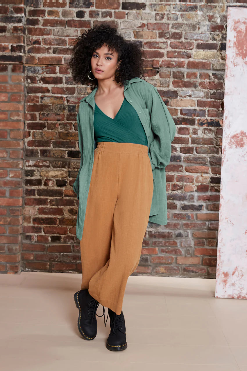 A woman wearing the Kurt Pants by Melow in Honey, featuring wide cropped legs, a flat-front elastic waist band, and slant pockets. She is wearing them with a green top and standing in front of a brick wall.