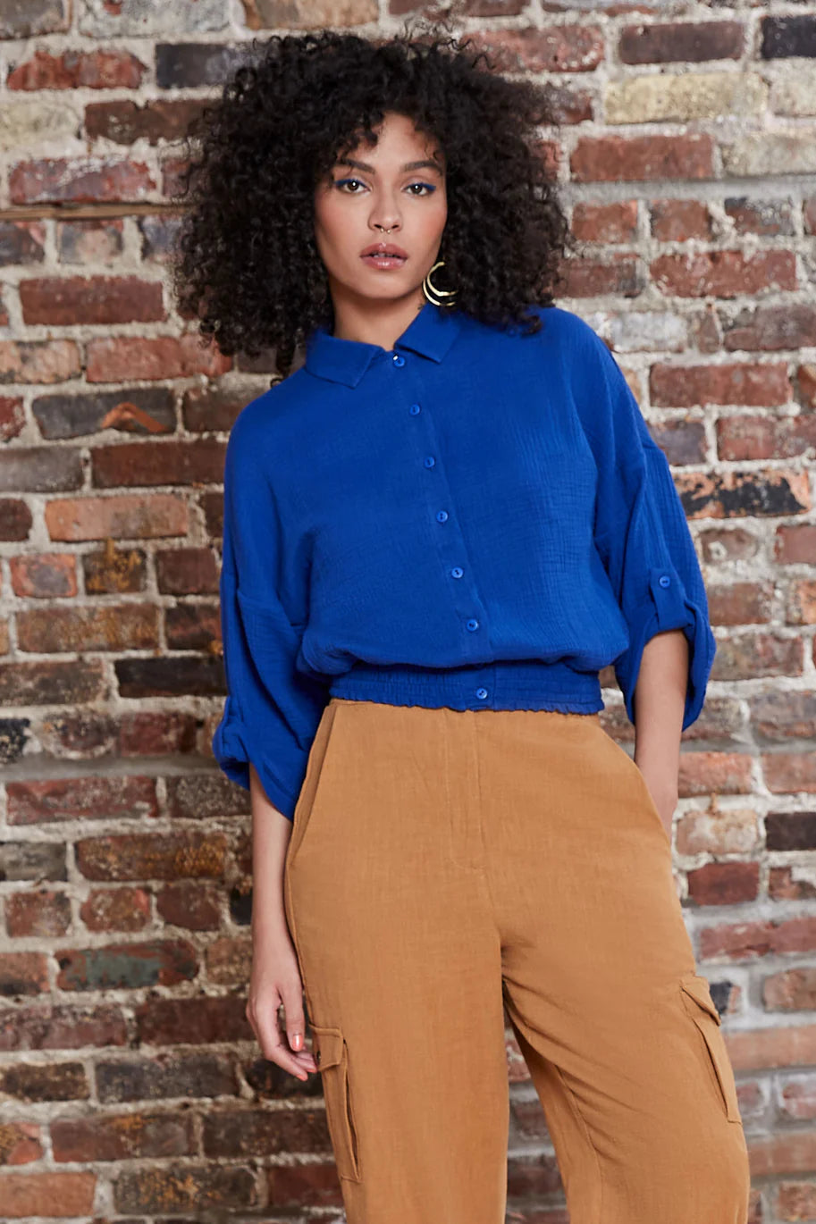 A woman wearing the Kemet Shirt by Melow in Cobalt, featuring a blouse collar, button front, honeycomb elastic waist, and cropped sleeves that fold and fasten with a button tab. She is wearing it with tan pants and standing in front of a brick wall.