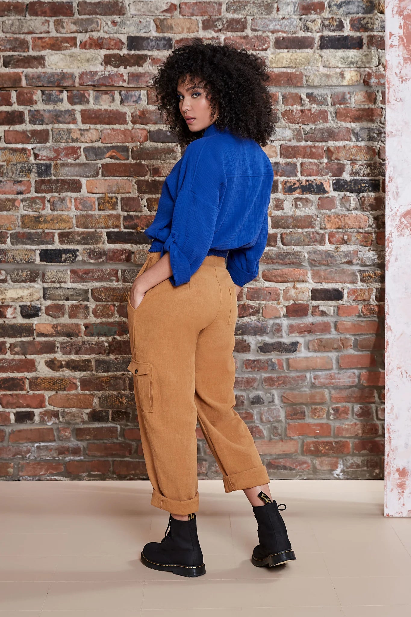 Back view of a woman wearing the Kemet Shirt by Melow in Cobalt, featuring a blouse collar, button front, honeycomb elastic waist, and cropped sleeves that fold and fasten with a button tab. She is wearing it with tan pants and standing in front of a brick wall.