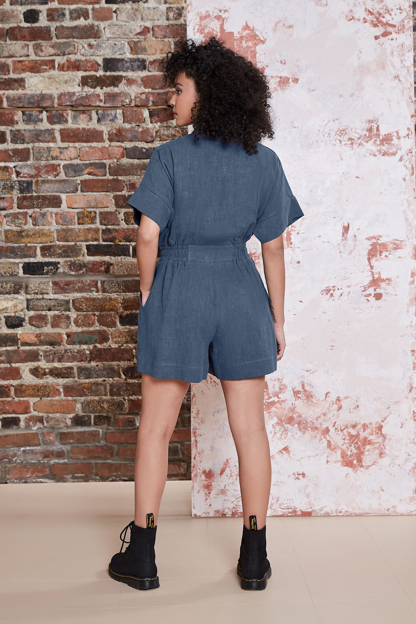 Back view of a woman wearing the Kerouac Jumpsuit by Melow in Slate, a short jumpsuit with a collar, zip front, chest pockets, elastic waist, and short sleeves. She is standing in front of a brick wall.