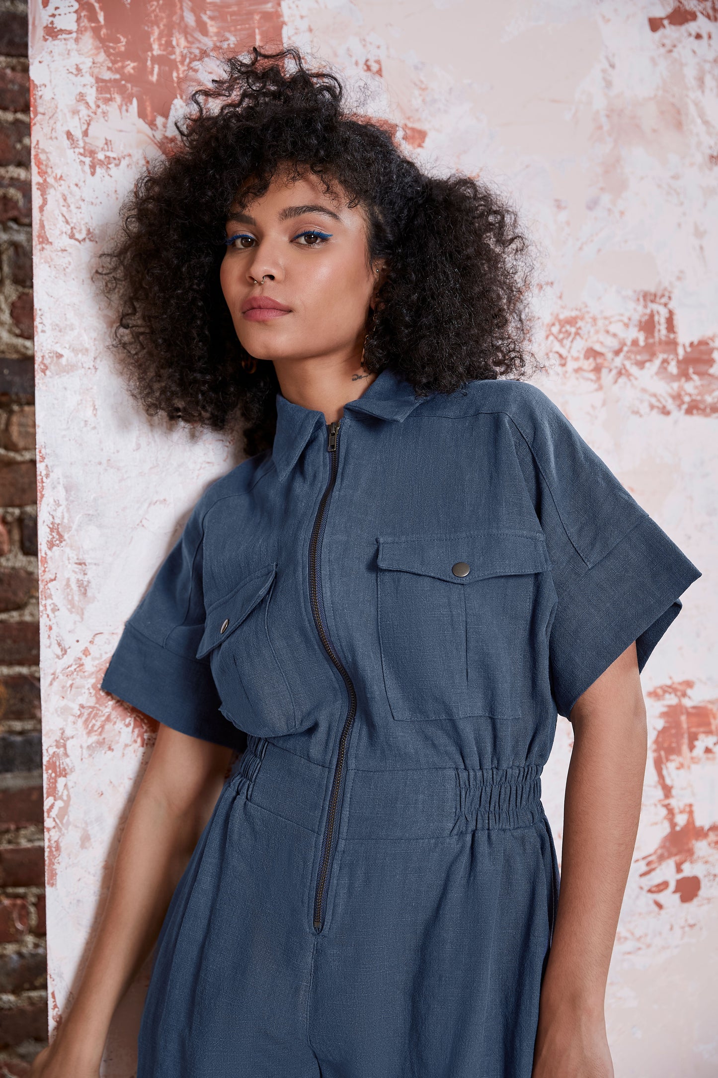 Waist up view of a woman wearing the Kerouac Jumpsuit by Melow in Slate, a short jumpsuit with a collar, zip front, chest pockets, elastic waist, and short sleeves. She is standing in front of a brick wall.