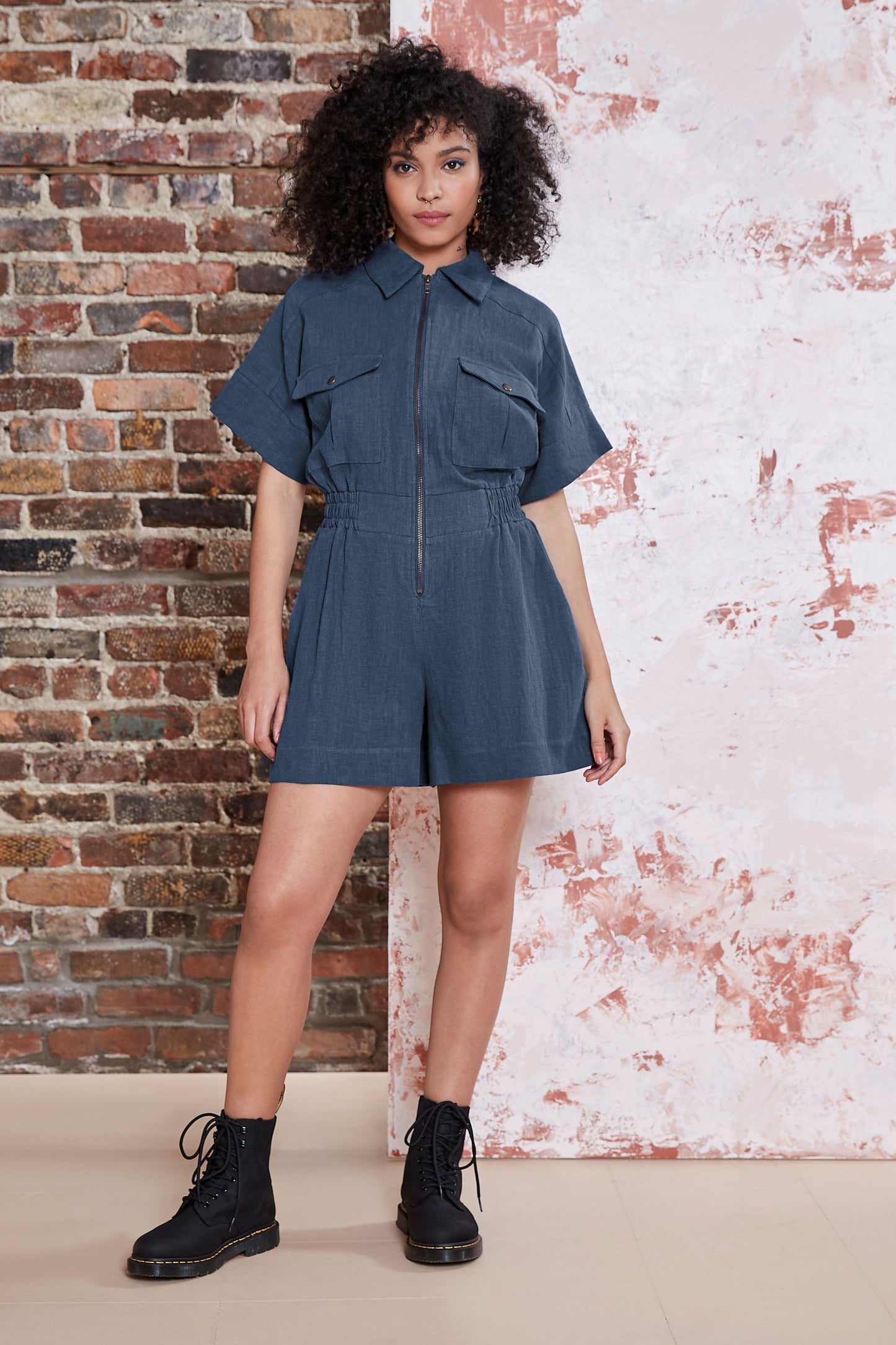 A woman wearing the Kerouac Jumpsuit by Melow in Slate, a short jumpsuit with a collar, zip front, chest pockets, elastic waist, and short sleeves. She is standing in front of a brick wall.