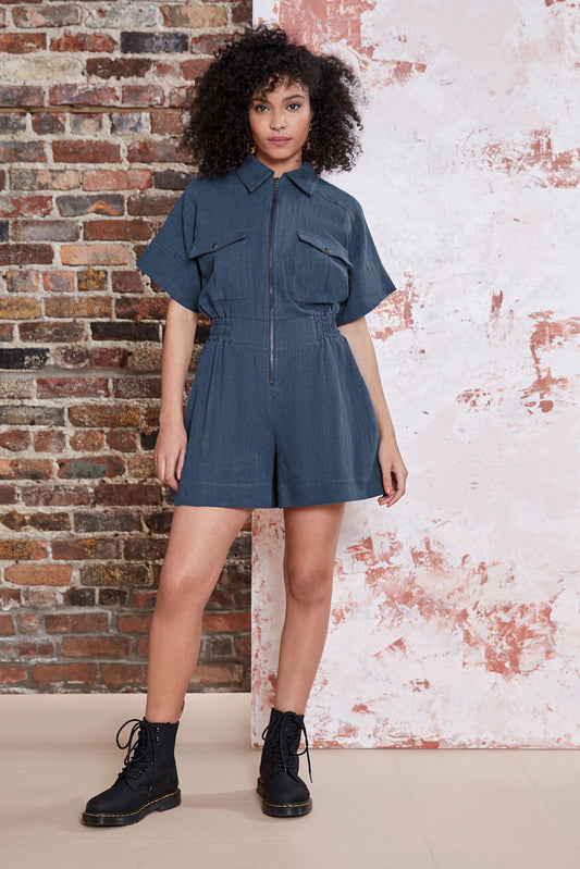 A woman wearing the Kerouac Jumpsuit by Melow in Slate, a short jumpsuit with a collar, zip front, chest pockets, elastic waist, and short sleeves. She is standing in front of a brick wall.