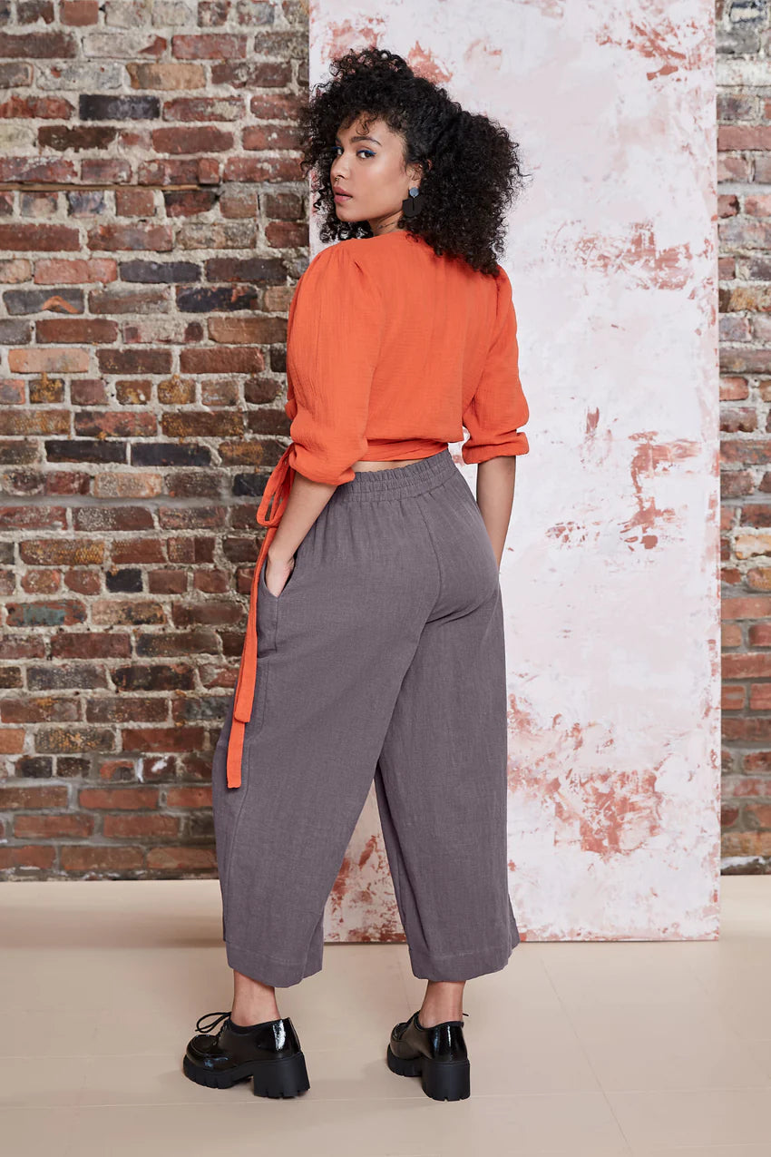 Back view of A woman wearing the Kurt Pants by Melow in Slate, featuring wide cropped legs, a flat-front elastic waist band, and slant pockets. She is wearing them with an orange top and standing in front of a brick wall.