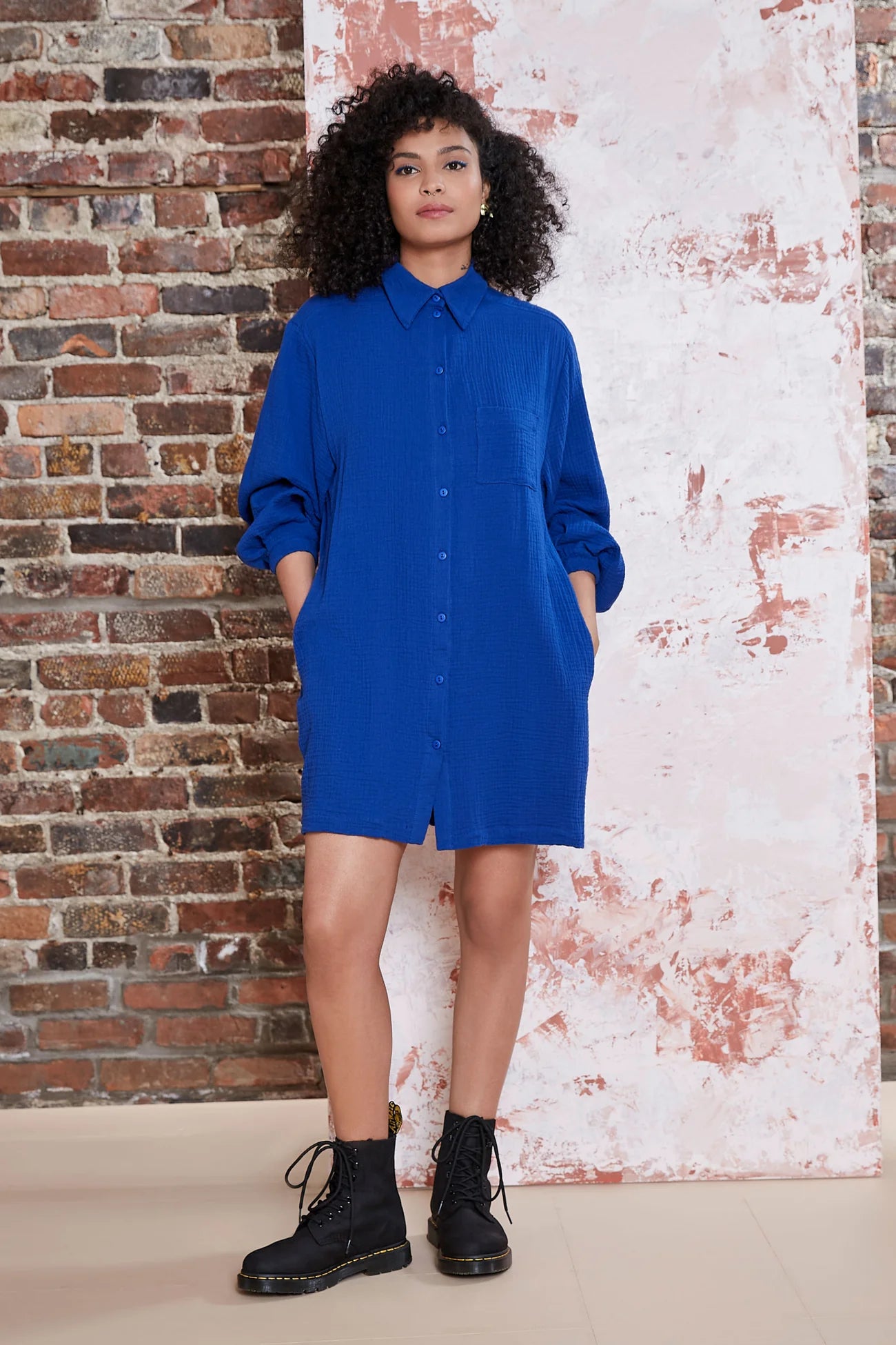 A woman wearing the Kronos Tunic by Melow in Colbalt, with a blouse collar, button front, an above-the-knee length and long sleeves that puff and gather at the wrists. She is standing in front of a brick wall.
