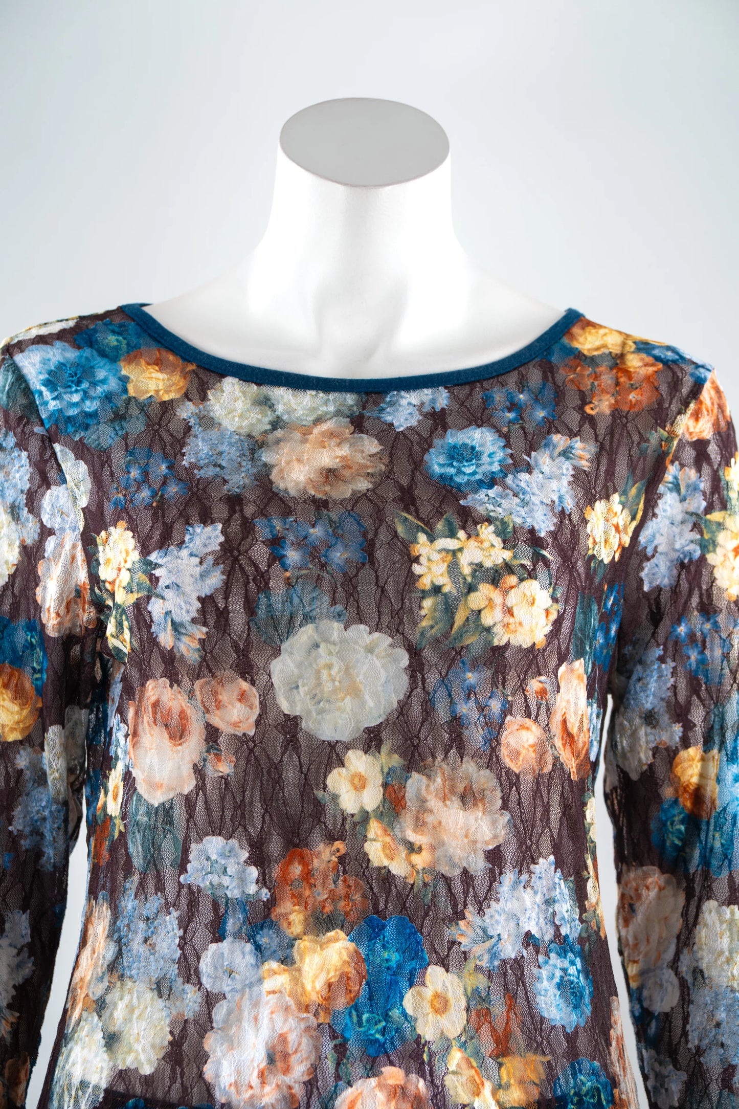 A close up view of the Aubade Lace Top by Karkass, featuring a floral print on a brown background, round neck, and long sleeves with contrasting teal at the collar, cuffs, and hem, is shown on a mannequin front of a white background.