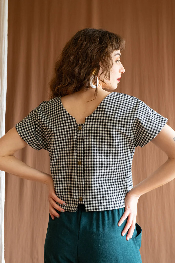 Back 3/4 view of a woman wearing the Brendola Top by Kazak in Black and White Vichy, a reversible top with a v-neck and snap front on one side and a round neck on the other side. It has short extended sleeves and a relaxed, cropped fit. She is wearing it with the Padoua Pants in Forest and standing in front of a brown curtain.