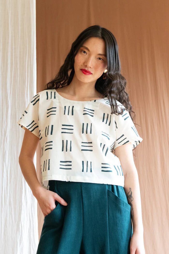 3/4 view of a woman wearing the Brendola Top by Kazak in Cream Lines, a reversible top with a v-neck and snap front on one side and a round neck on the other side. It has short extended sleeves and a relaxed, cropped fit. She is wearing it with the Padoua Pants in Forest and standing in front of a brown curtain.
