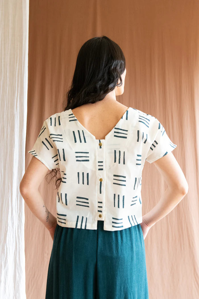 Back 3/4 view of a woman wearing the Brendola Top by Kazak in Cream Lines, a reversible top with a v-neck and snap front on one side and a round neck on the other side. It has short extended sleeves and a relaxed, cropped fit. She is wearing it with the Padoua Pants in Forest and standing in front of a brown curtain.