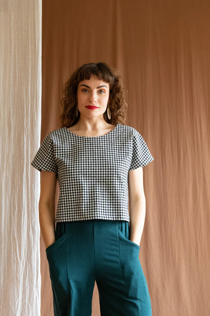 3/4 view of a woman wearing the Brendola Top by Kazak in Black and White Vichy, a reversible top with a v-neck and snap front on one side and a round neck on the other side. It has short extended sleeves and a relaxed, cropped fit. She is wearing it with the Padoua Pants in Forest and standing in front of a brown curtain.