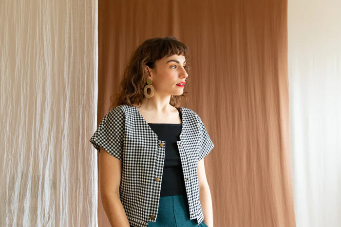 Waist up view of a woman wearing the Brendola Top by Kazak in Black and White Vichy, a reversible top with a v-neck and snap front on one side and a round neck on the other side. It has short extended sleeves and a relaxed, cropped fit. She is wearing it with the Padoua Pants in Forest and standing in front of a brown curtain.