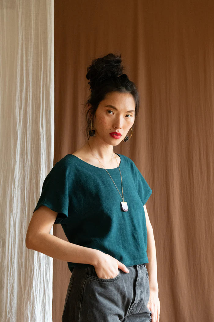 3/4 view of a woman wearing the Brendola Top by Kazak in Forest Green, a reversible top with a v-neck and snap front on one side and a round neck on the other side. It has short extended sleeves and a relaxed, cropped fit. She is wearing it with black jeans and standing in front of a brown curtain.