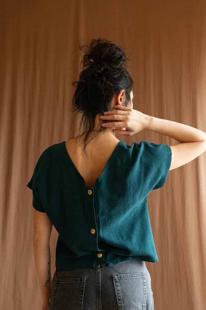 3/4 view of a woman wearing the Brendola Top by Kazak in Forest Green, a reversible top with a v-neck and snap front on one side and a round neck on the other side. It has short extended sleeves and a relaxed, cropped fit. She is wearing it with black jeans and standing in front of a brown curtain.