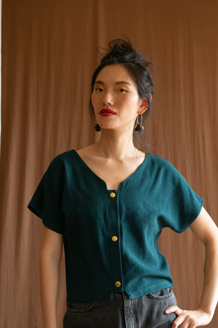 3/4 view of a woman wearing the Brendola Top by Kazak in Forest Green, a reversible top with a v-neck and snap front on one side and a round neck on the other side. It has short extended sleeves and a relaxed, cropped fit. She is wearing it with black jeans and standing in front of a brown curtain.