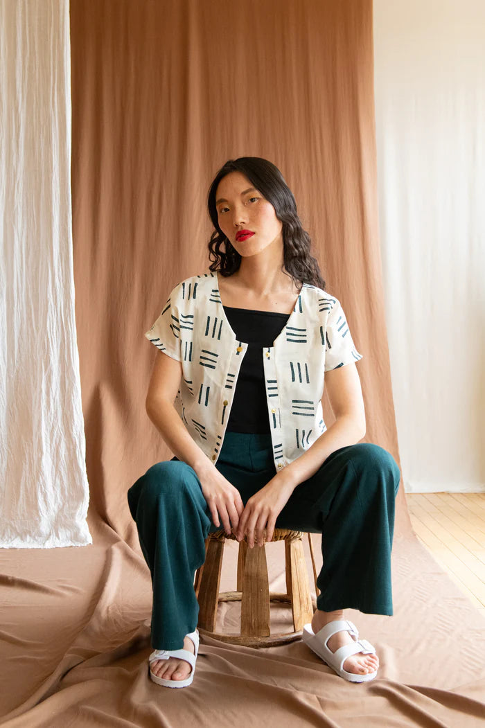 A woman wearing the Brendola Top by Kazak in Cream Lines, a reversible top with a v-neck and snap front on one side and a round neck on the other side. It has short extended sleeves and a relaxed, cropped fit. She is wearing it with the Padoua Pants in Forest and sitting in front of a brown curtain.