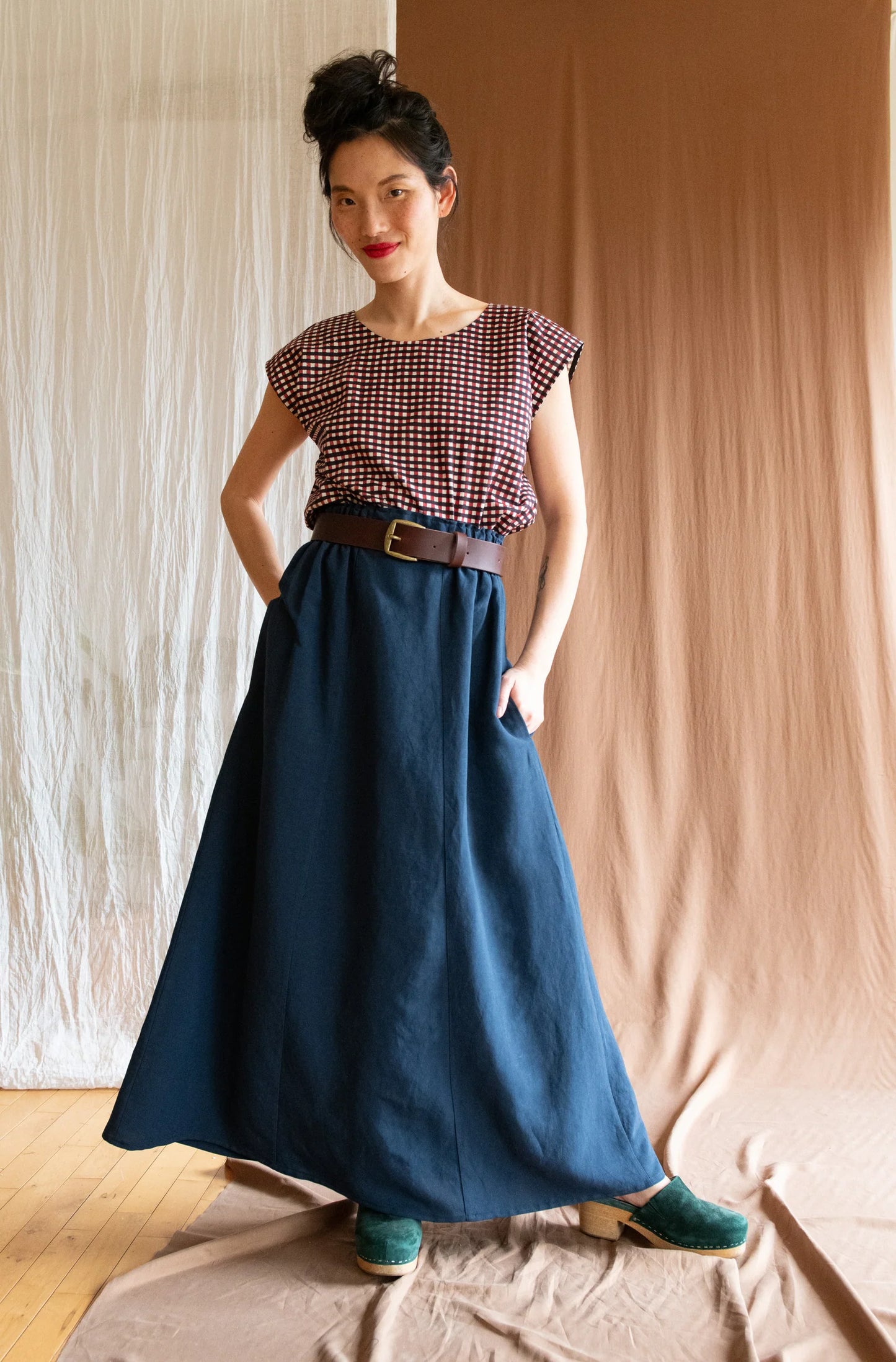 A woman wearing the Sandrigo Top by Kazak in Charcoal and Red Stamped Checks, with short extended sleeves, a round and wide neck, and a loose fit. She is wearing it with the Verona Skirt in Indigo and standing in front of a brown curtain.