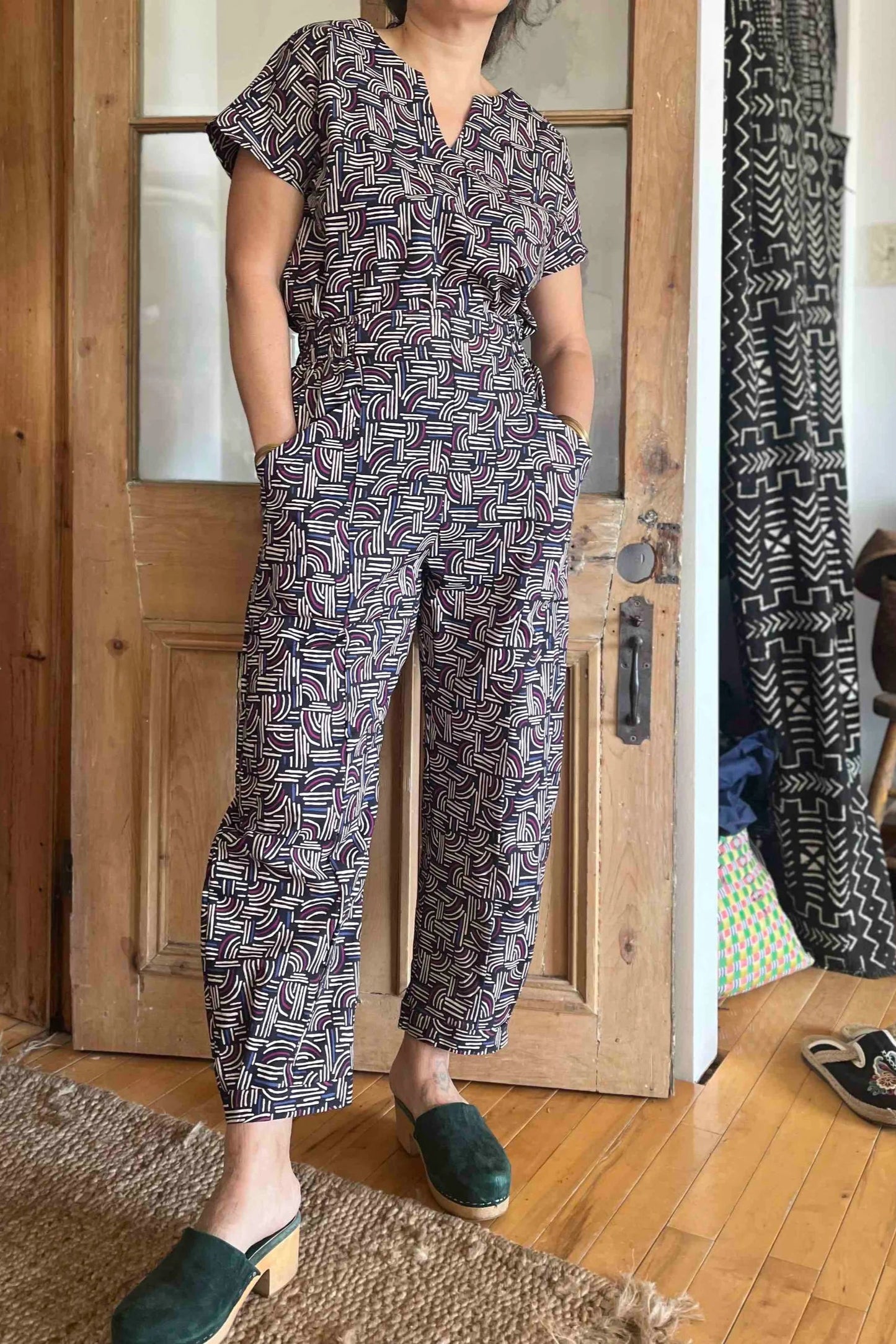 Neck down view of a woman wearing the Lucca Top by Kazak in Night Basketwork print, a short sleeved to with a notched V-neckline and a pleated yoke at the back. She is wearing it with the matching Sienna Pants and is standing in front of a wooden door.