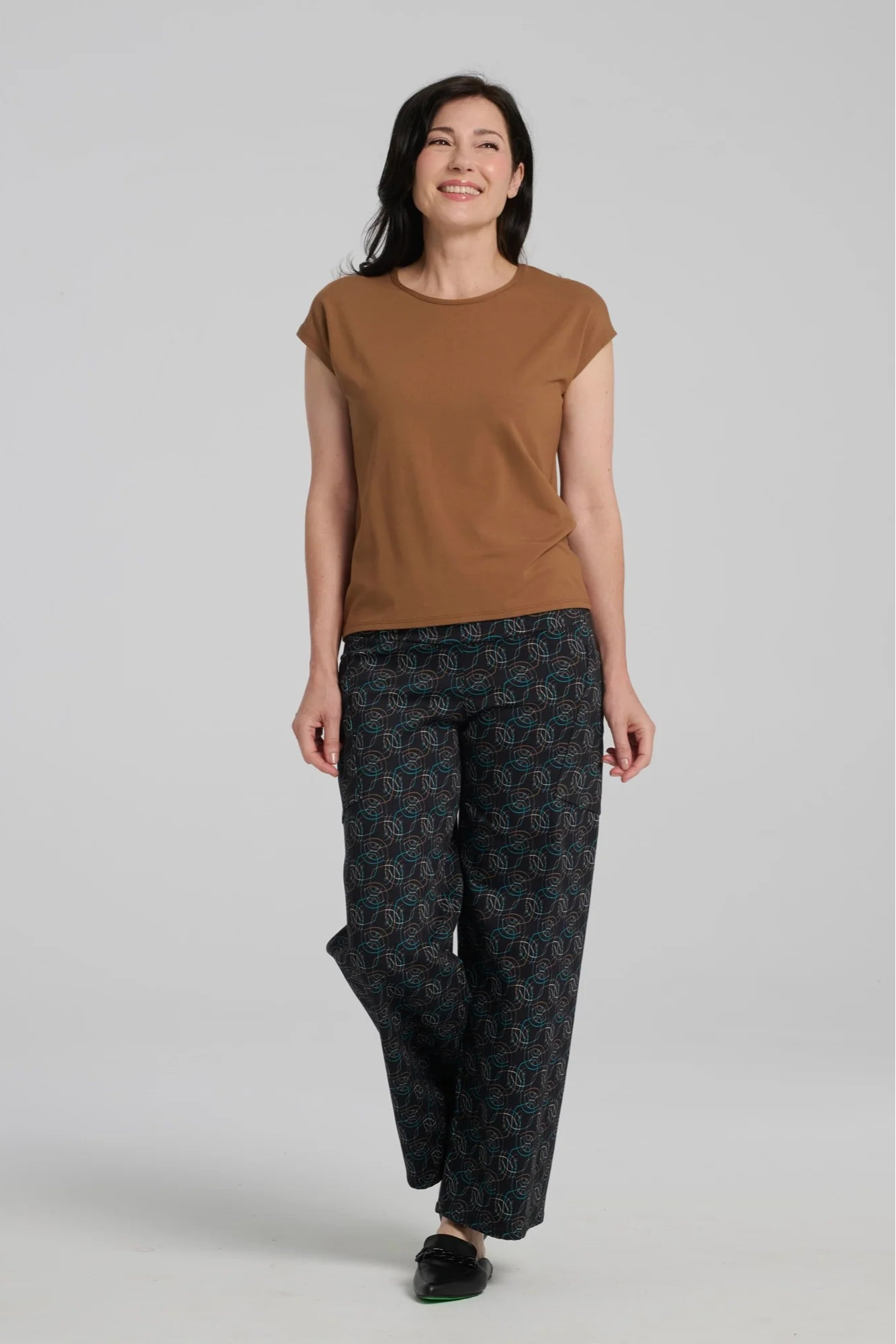 A woman wearing the Elma Pants by Kollontai in Turquoise on black print, straight legged pants with a wide elastic waist and large slant pockets. She is wearing them with the Eowyn Top in Caramel and standing in front of a grey background.