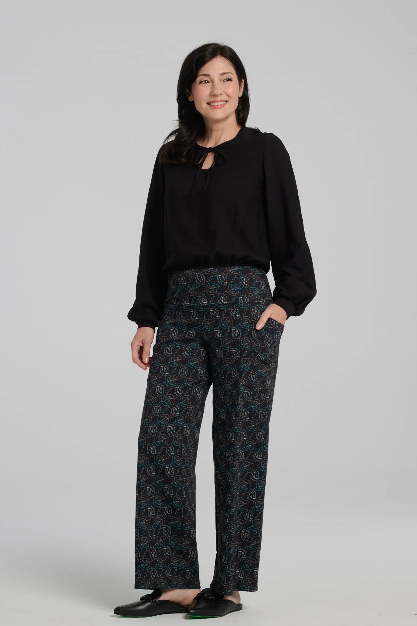 A woman wearing the Elma Pants by Kollontai in Turquoise on black print, straight legged pants with a wide elastic waist and large slant pockets. She is wearing them with a black top and standing in front of a grey background.