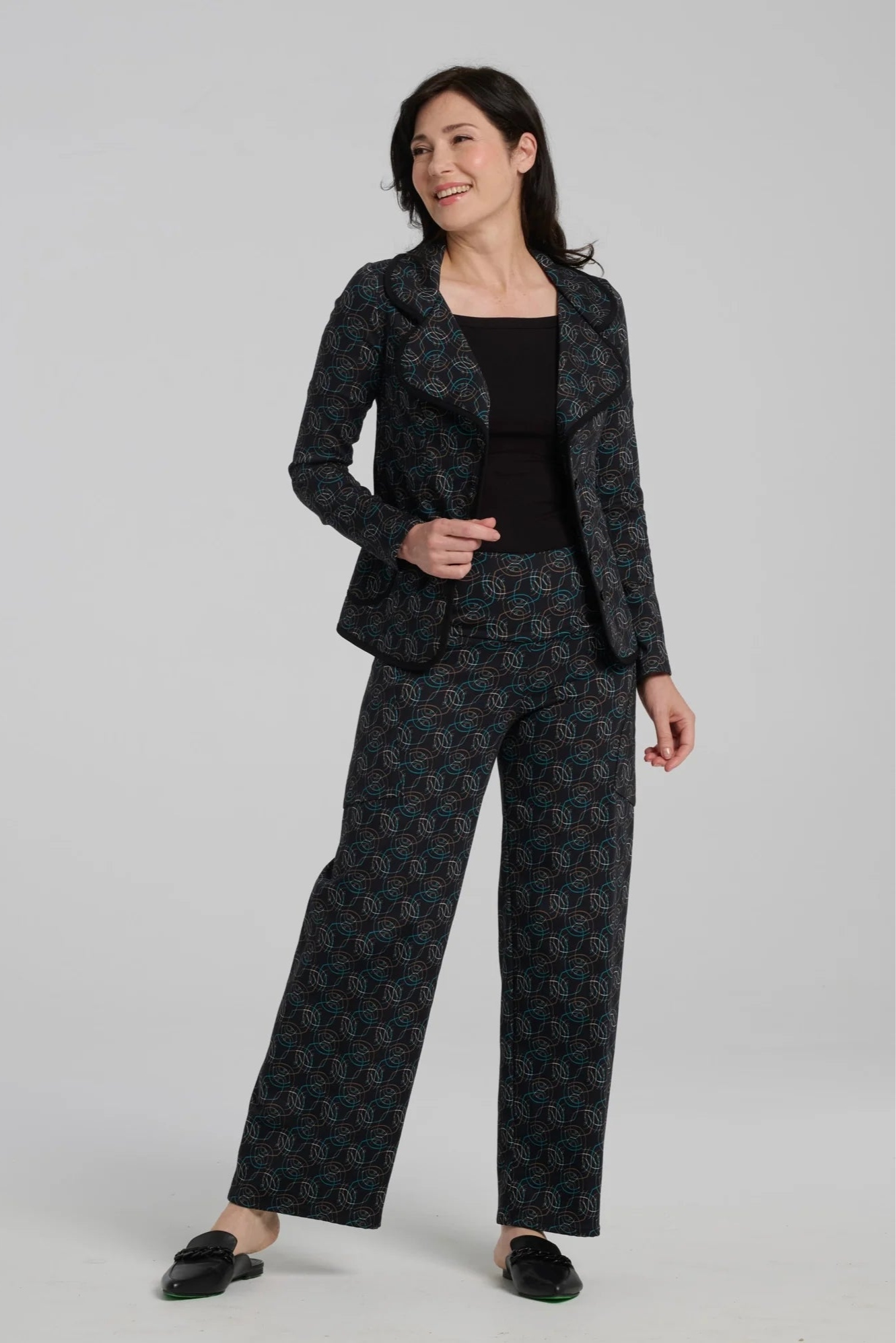 A woman wearing the Elma Pants by Kollontai in Turquoise on black print, straight legged pants with a wide elatic waist and large slant pockets. She is wearing them with a black top and matching jacket and standing in front of a grey background.