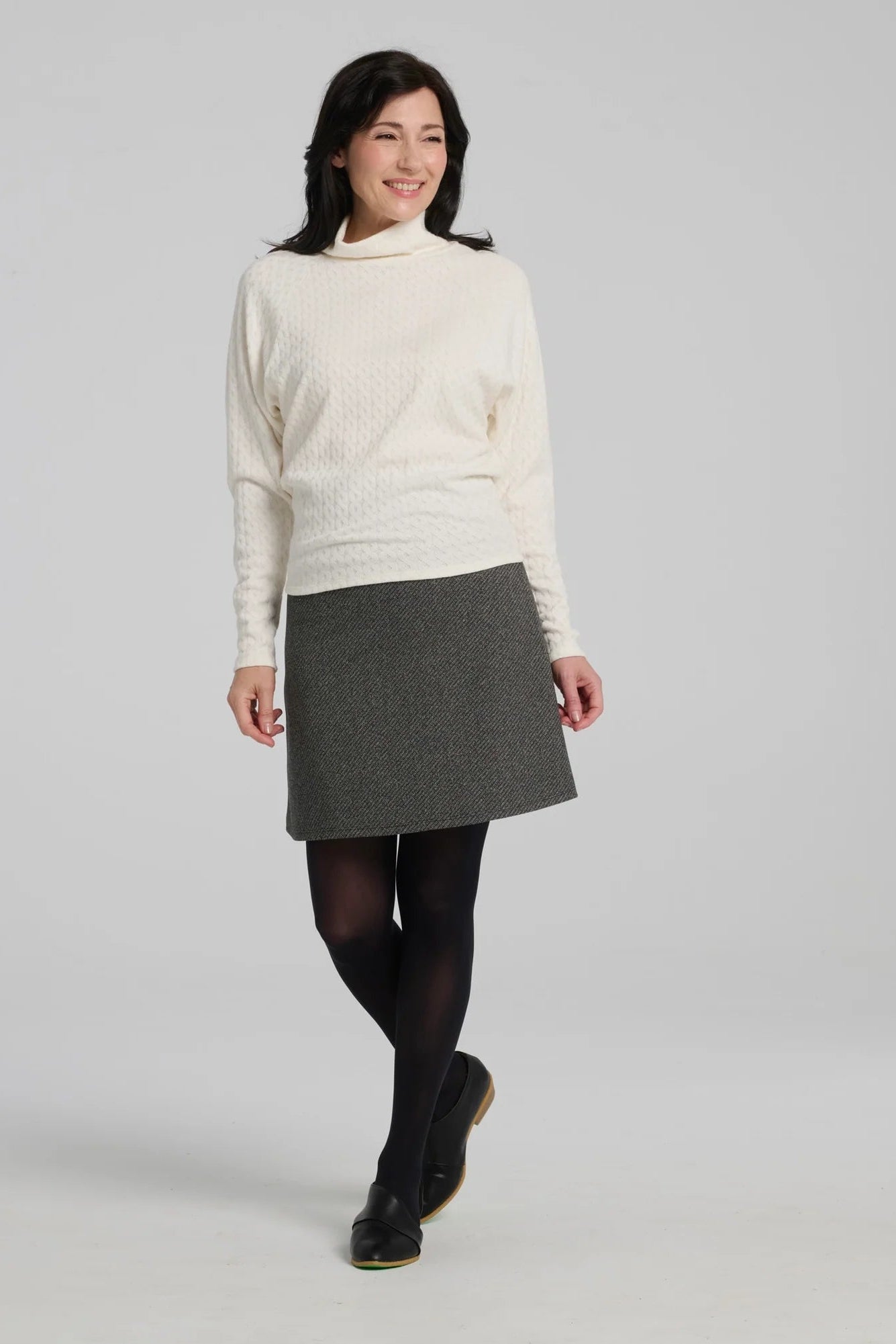 A woman wearing the Frost Skirt by Kollontai in Charcoal tweed, an above the knee length A-line skirt with an elastic waist. She is wearing it with the Plume Top in White and standing in front of a grey background.