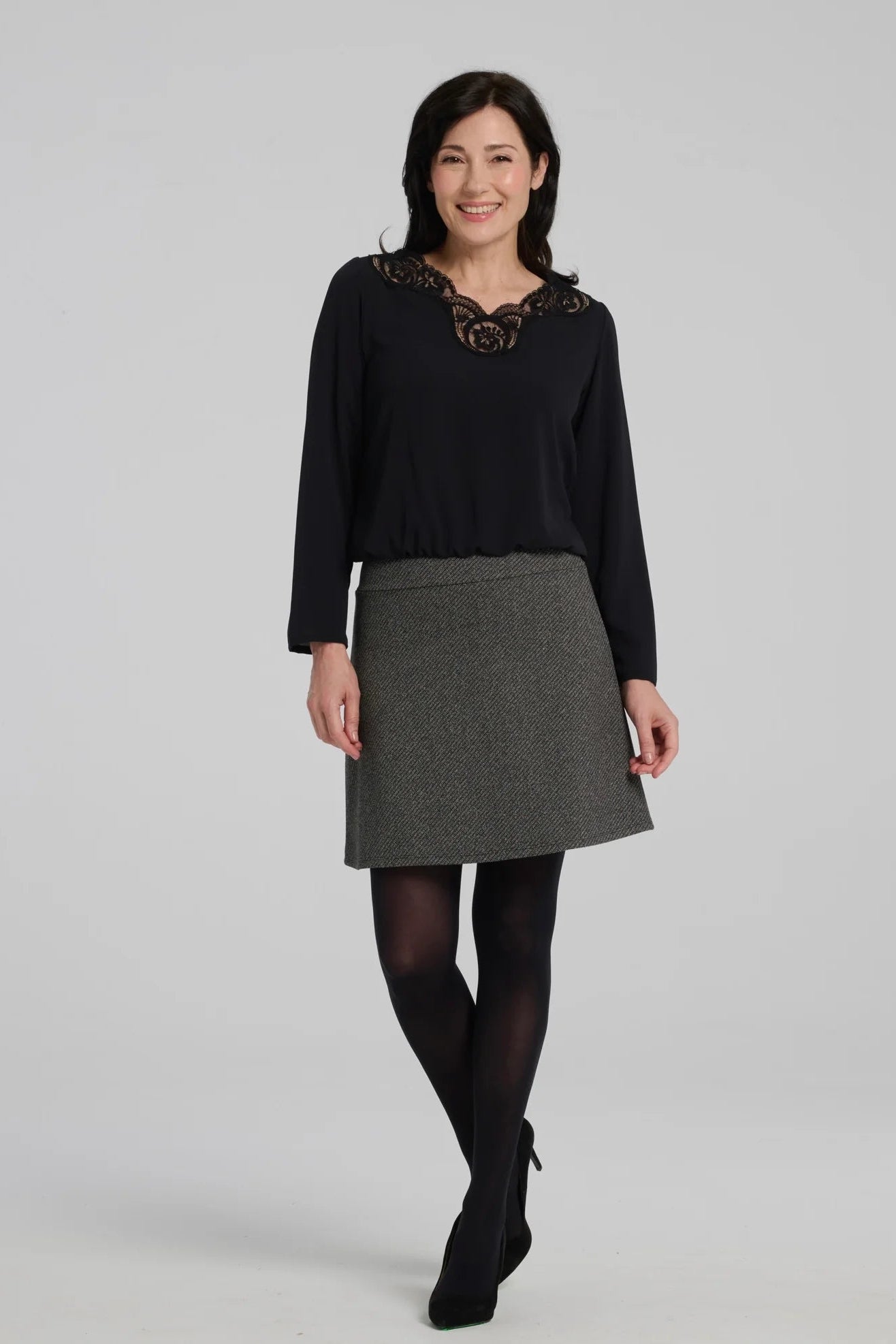 A woman wearing the Frost Skirt by Kollontai in Charcoal tweed, an above the knee length A-line skirt with an elastic waist. She is wearing it with a black top and standing in front of a grey background.