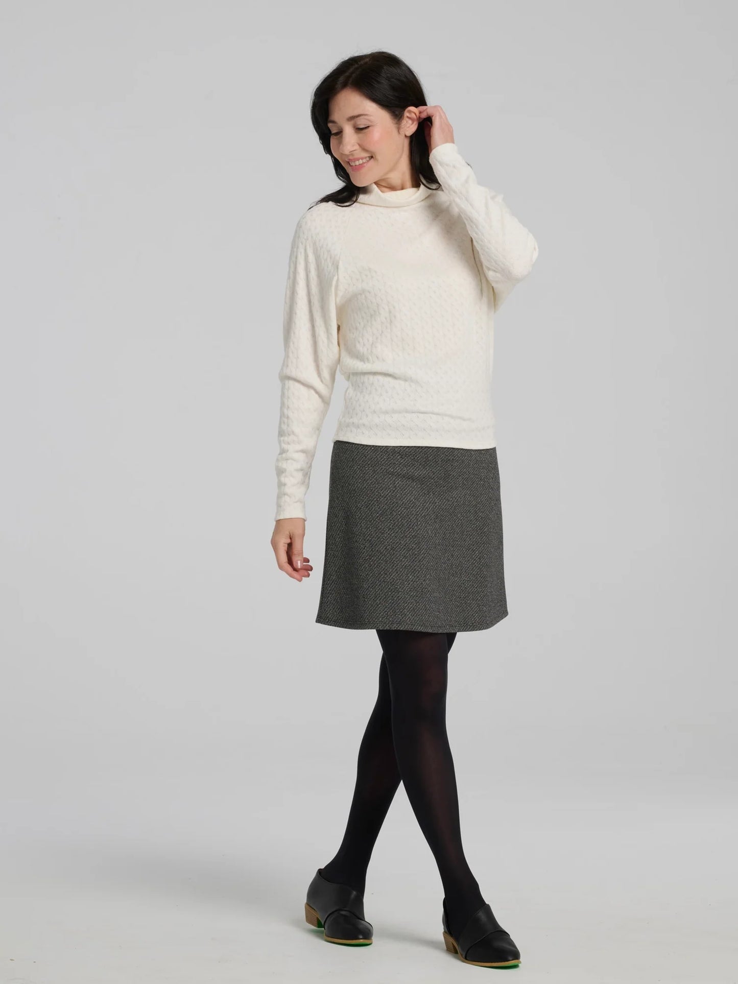 A woman wearing the Frost Skirt by Kollontai in Charcoal tweed, an above the knee length A-line skirt with an elastic waist. She is wearing it with a black top and standing in front of a grey background.
