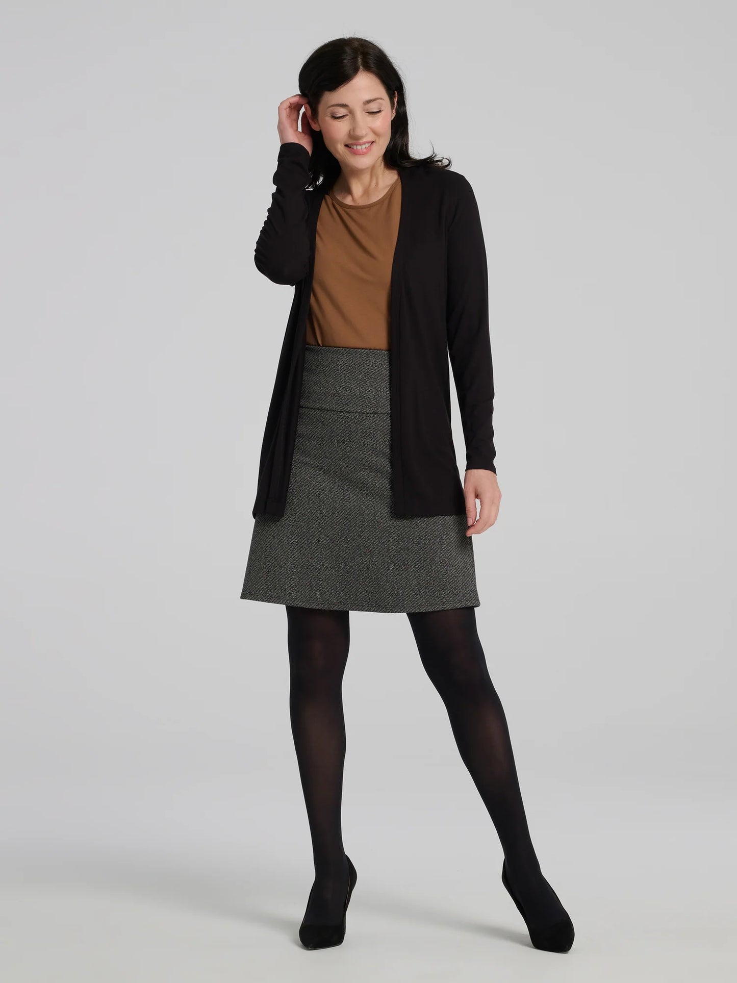A woman wearing the Frost Skirt by Kollontai in Charcoal tweed, an above the knee length A-line skirt with an elastic waist. She is wearing it with a brown top and black cardigan top and standing in front of a grey background.