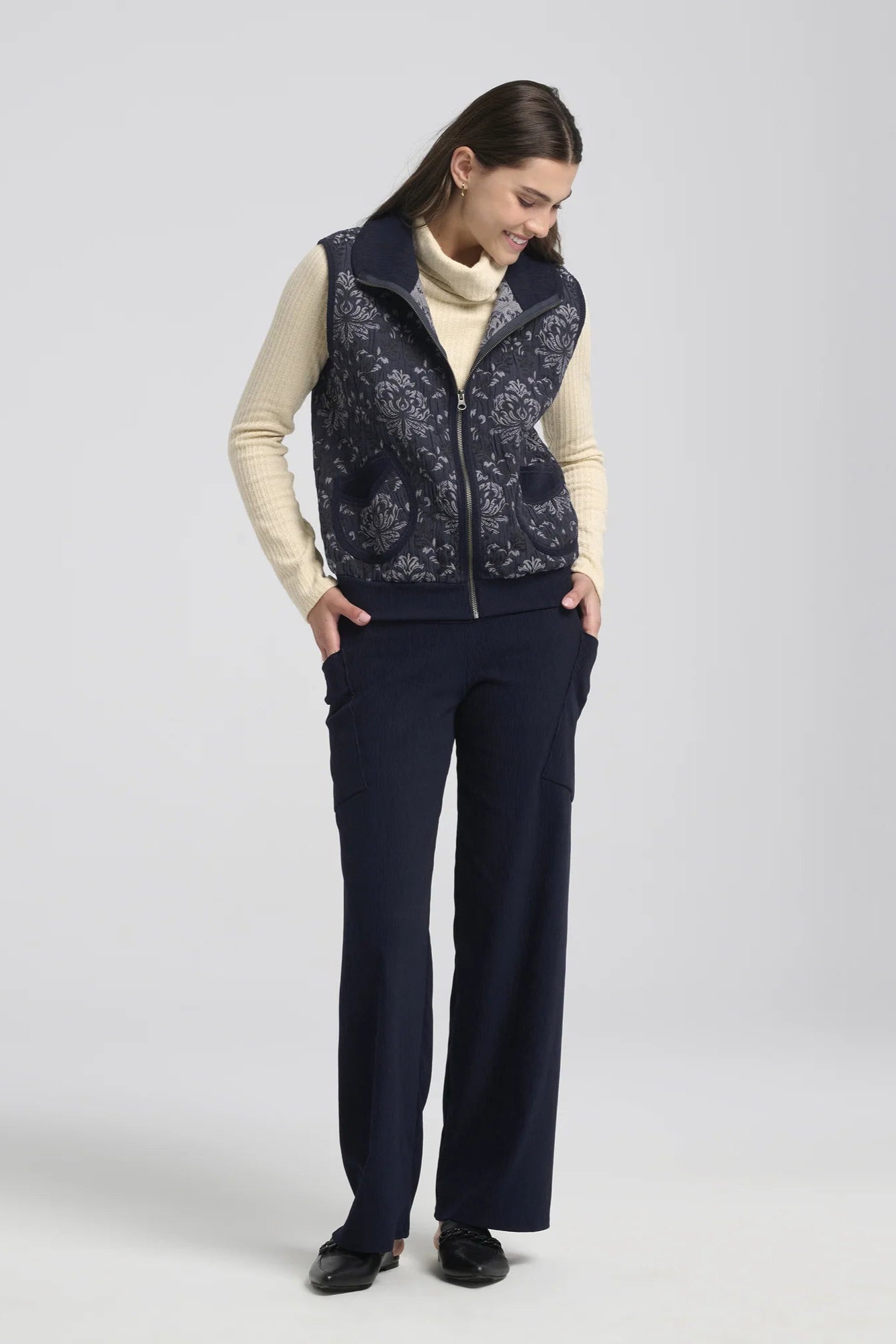 A woman wearing the Hermione Pants by Kollontai in Navy, stretchy corduroy pants with a wide pull-one waistband, wide legs, and angled patch pockets. She is wearing them with a cream sweater and a vest and standing in front of a grey background.
