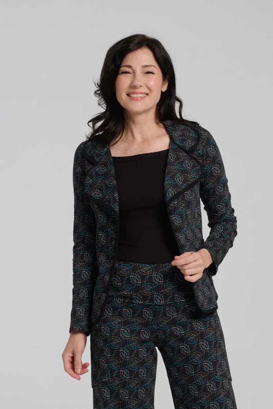 A woman wearing the Nara Jacket by Kollontai in Turquoise, a blazer with a turquoise on black geometric print, wit a rounded lapel collar, slim fit, and side pockets. She is wearing it with a black top and the matching Elma Pant and standing in front of a grey background.