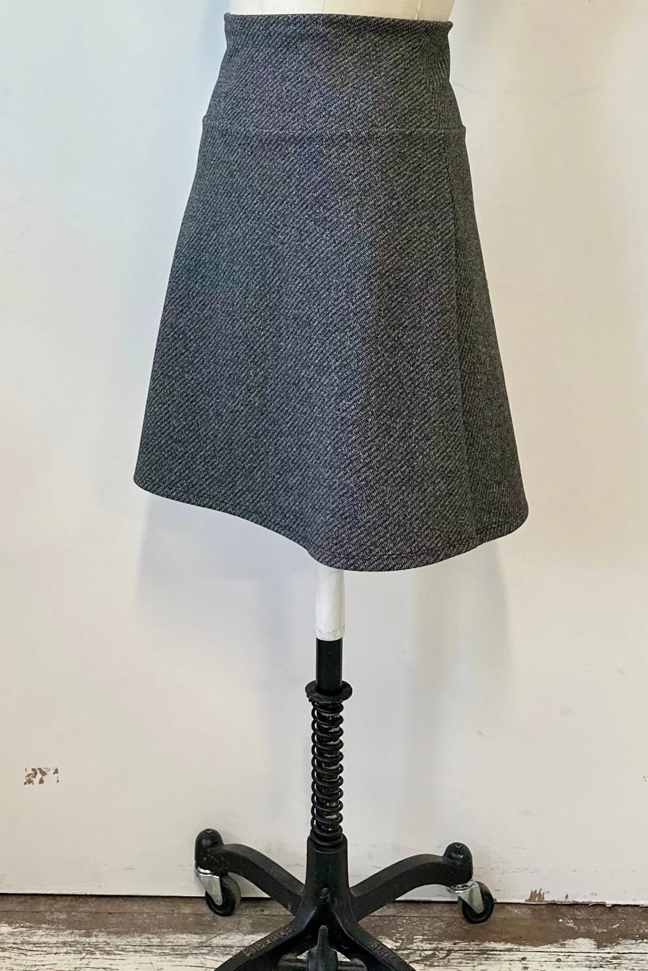 A side view of the Frost Skirt by Kollontai in Charcoal tweed, an above the knee length A-line skirt with an elastic waist, is shown on a mannequin in front of a white wall.