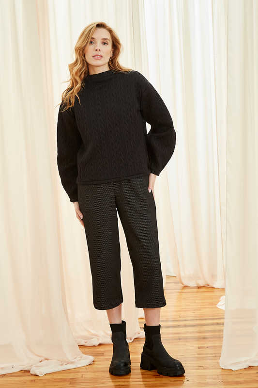 A woman wearing the Lucas Pants by Melow in Black, featuring a diagonal stripe texture, a high elastic waist, front pleats, and 3/4 length legs. She is wearing them with a black sweater and  standing in front of a white curtain.