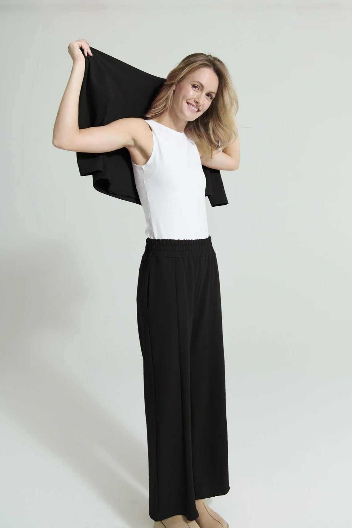 A woman wearing the Romaine Sweater and Tadoussac Pants by Message Factory in Black: a sweatshirt with a funnel neck and long drop sleeves and flared sweatpants with an elastic waist, both in a bamboo/cotton blend. She is wearing a white tank top with them and is standing in front of a white background.