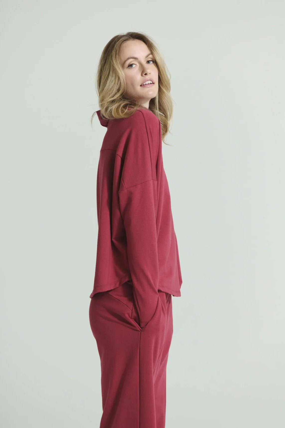 Side view of a woman wearing the Romaine Sweater by Message Factory in Raspberry, a bamboo/organic cotton sweatshirt with a funnel neck, a loose fit, and long drop sleeves. She is wearing the matching Tadoussac Pants and standing in front of a white background.