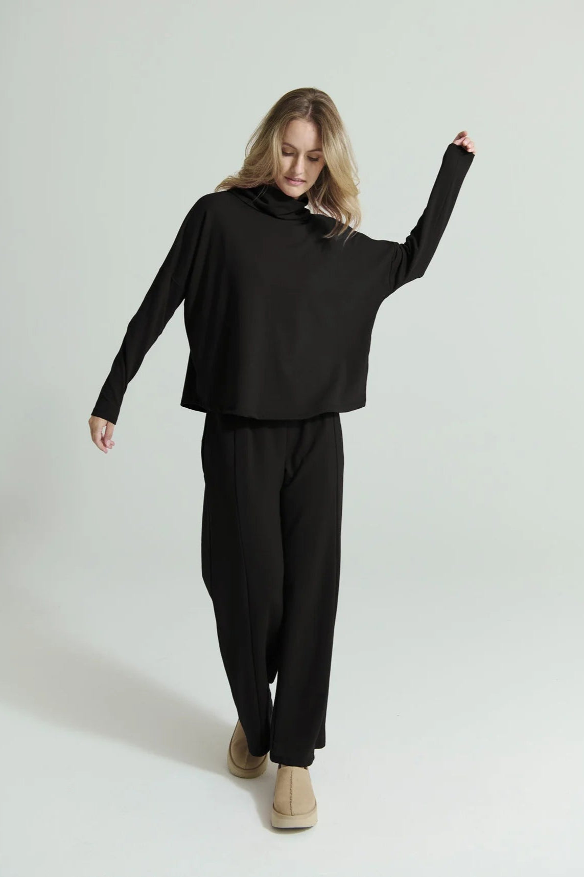 A woman wearing the Romaine Sweater by Message Factory in Black, a bamboo/organic cotton sweatshirt with a funnel neck , a loose fit, and long drop sleeves. She is wearing the matching Tadoussac Pants and standing in front of a white background.