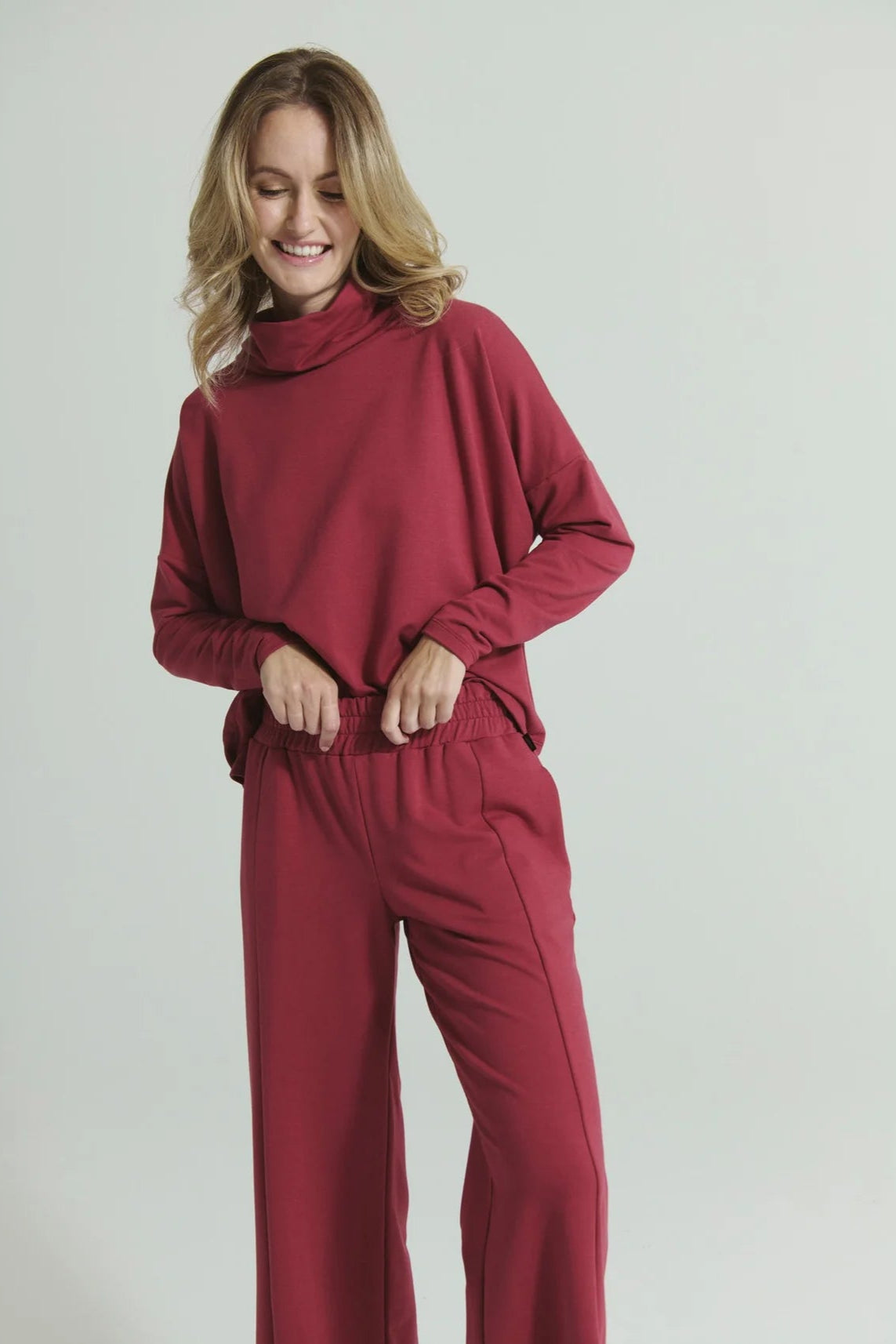 A woman wearing the Romaine Sweater and Tadoussac Pants by Message Factory in Raspberry: a sweatshirt with a funnel neck and long drop sleeves and flared sweatpants with an elastic waist, both in a bamboo/cotton blend. She is standing in front of a white background.