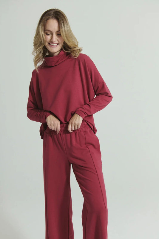 A woman wearing the Romaine Sweater and Tadoussac Pants by Message Factory in Raspberry: a sweatshirt with a funnel neck and long drop sleeves and flared sweatpants with an elastic waist, both in a bamboo/cotton blend. She is standing in front of a white background.