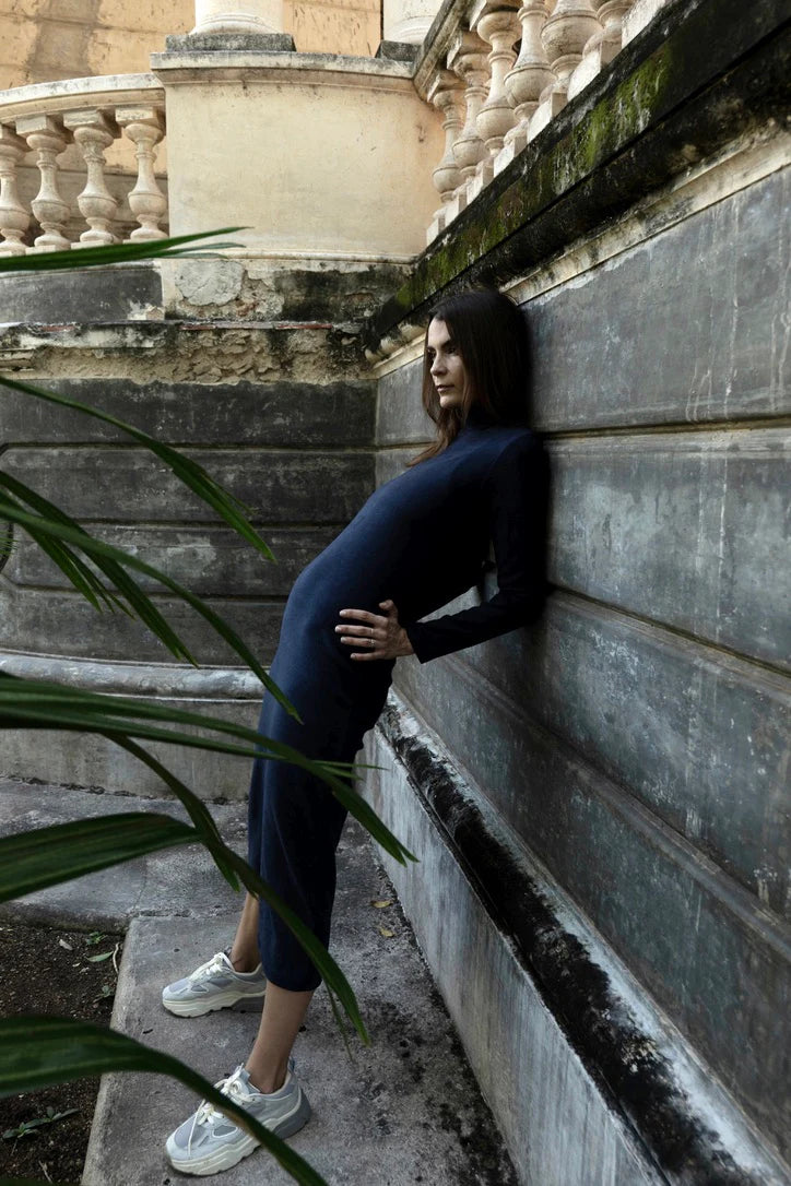 Woman in wearing the Mecca Dress by MAS in Blueberry, a long slim-fitting turtleneck dress. She is standing next to a stone wall.