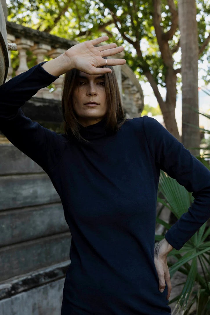Woman in wearing the Mecca Dress by MAS in Blueberry, a long slim-fitting turtleneck dress. She is standing next to a stone wall with greenery in the background.