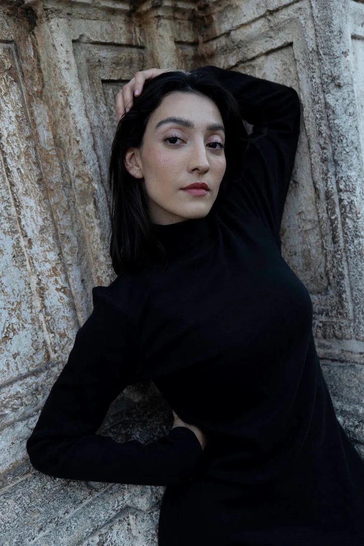 Woman in wearing the Mecca Dress by MAS in Black, a long slim-fitting turtleneck dress. She is standing next to a stone wall.
