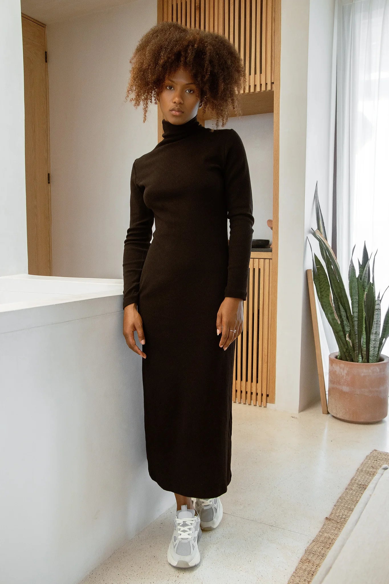 Woman in wearing the Mecca Dress by MAS in Pepper a long slim-fitting turtleneck dress. She is standing next to a white counter.