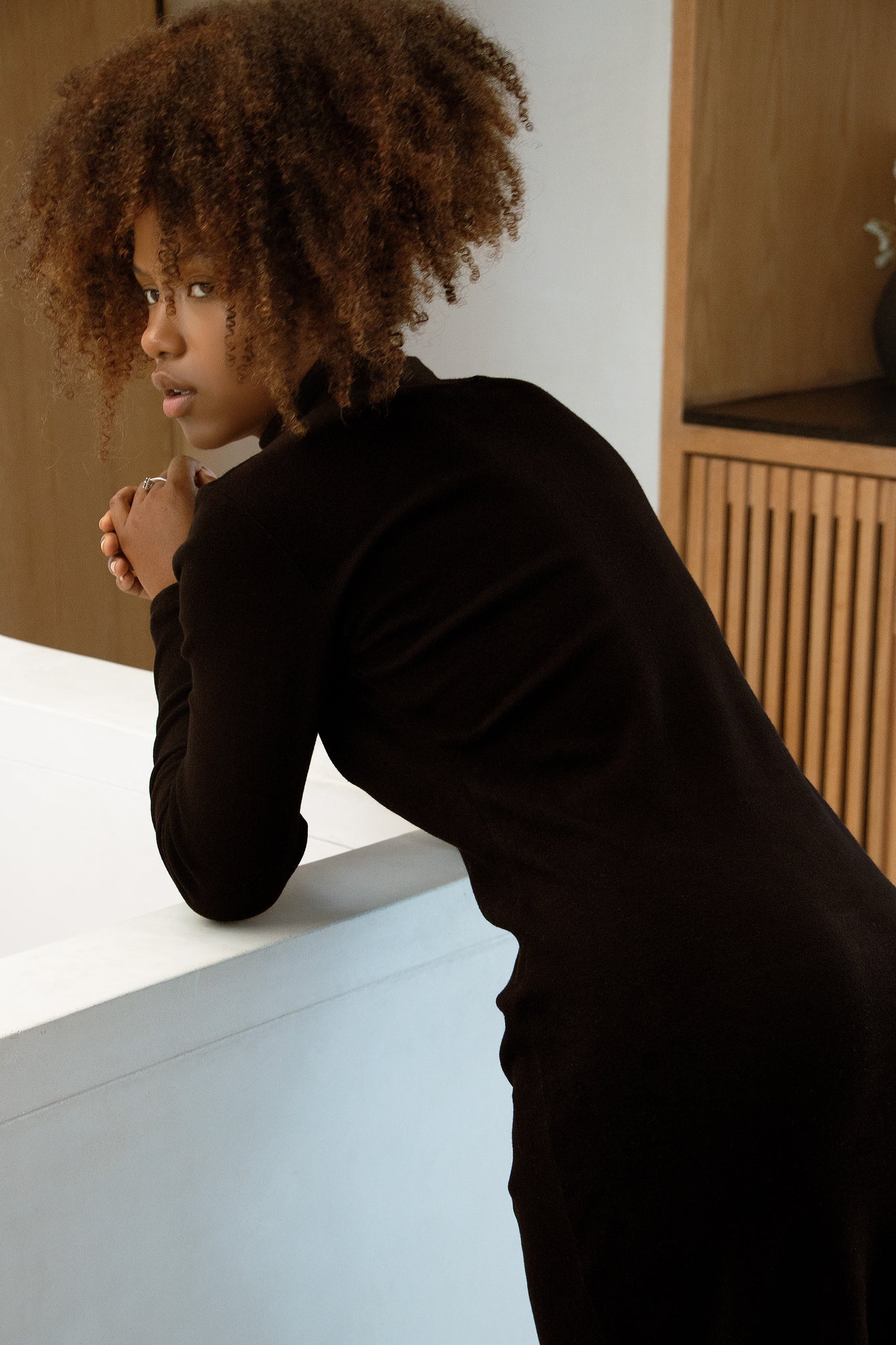 Back view of a woman in wearing the Mecca Dress by MAS in Pepper, a long slim-fitting turtleneck dress. She is leaning over a white countertop.