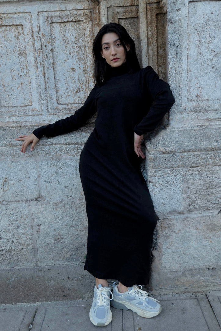 Woman in wearing the Mecca Dress by MAS in Pepper, a long slim-fitting turtleneck dress. She is standing next to a stone wall.