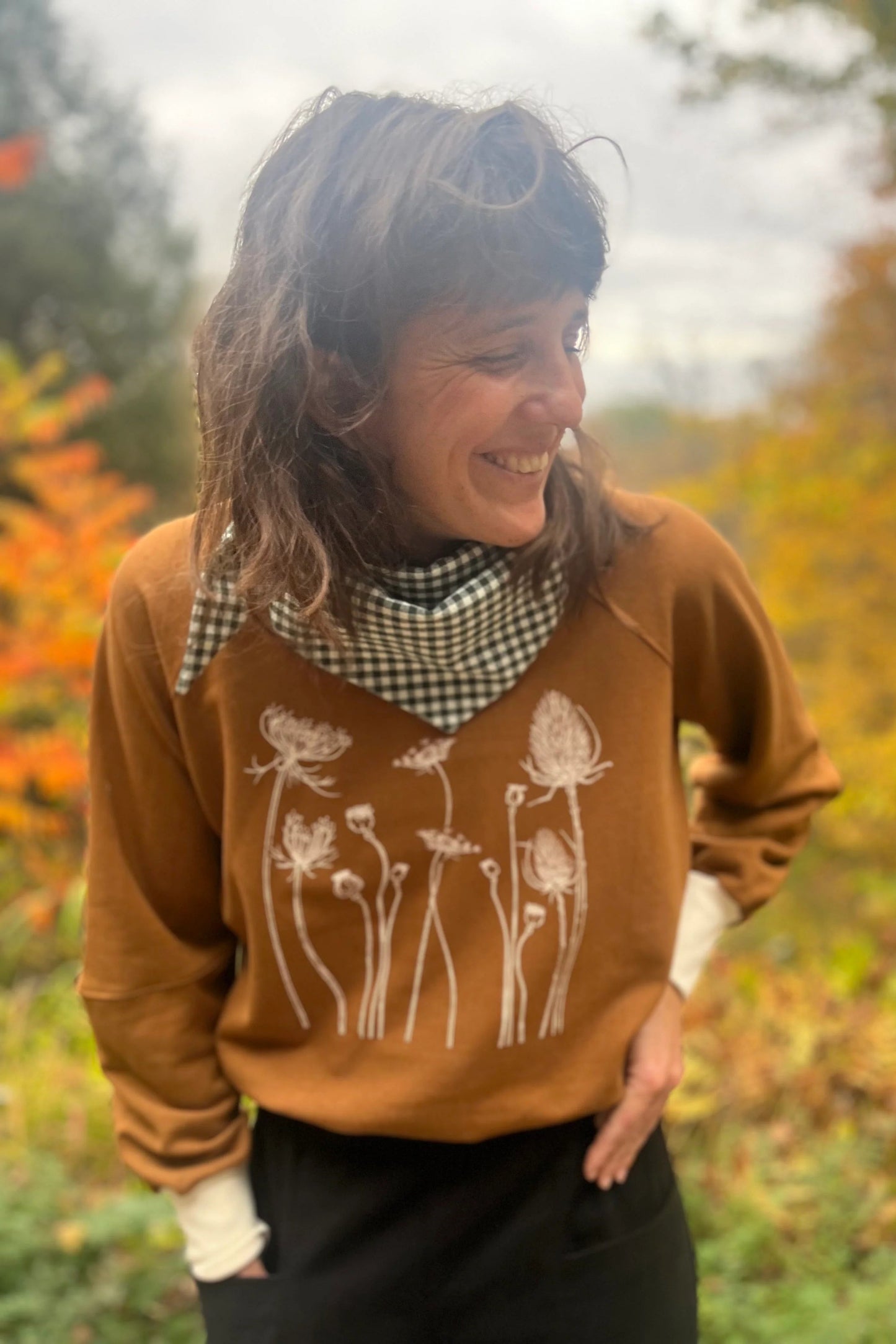 A woman wearing the Marilou Crewneck with Flowers by Kazak in Teak, a raglan sleeved sweatshirt with a screen print design. She is wearing it with jeans and standing in a garden.