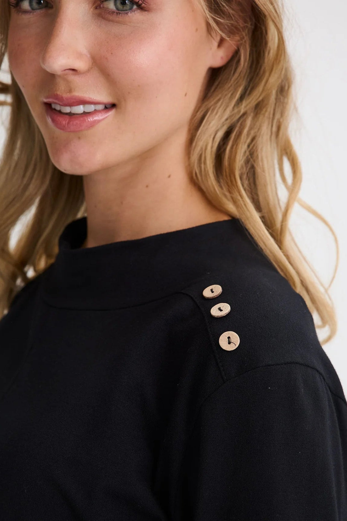 Close up view of the shoulder button detail on the Carleton Top by Message Factory in a Black, worn by a woman standing in front of a white background.